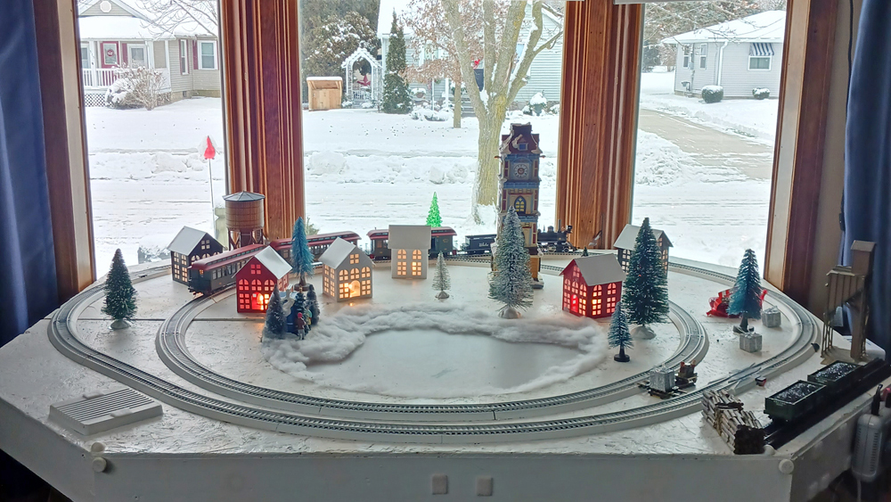 A model railroad layout with a Christmas theme is seen in a bay window of a home with snow on the ground outside