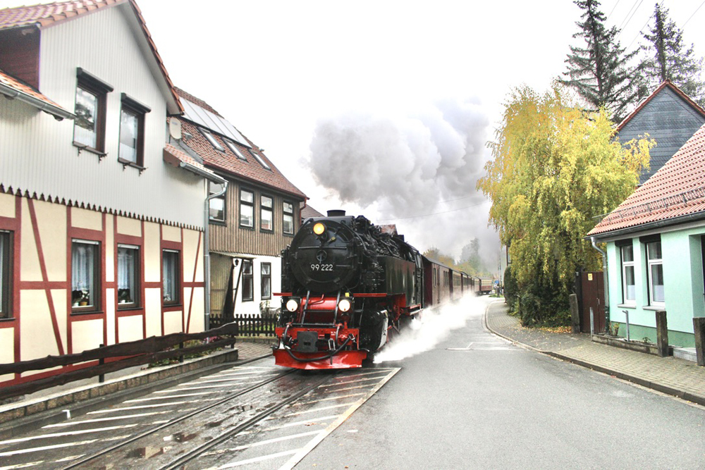 Steam-powered passenger train on narrow street with buildings on either side. 