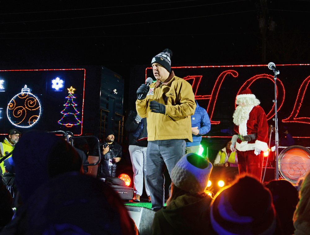 Man in mustard-yellow jacket speaking with Santa Claus in background