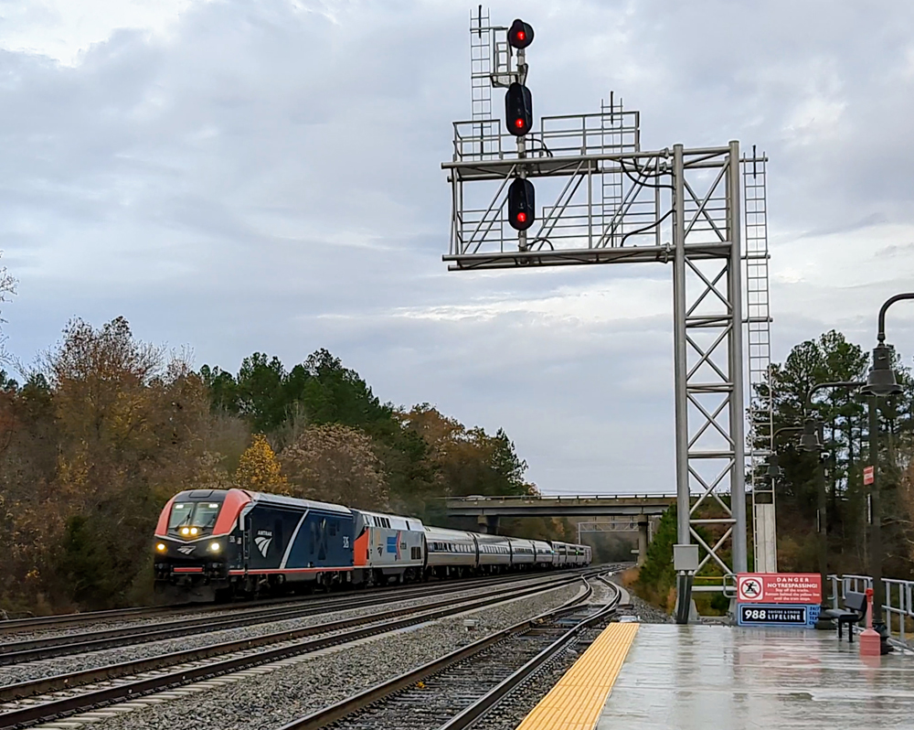 Passenger train with blue locomotive and heritage Amtrak locomotive passing next to signal bridge