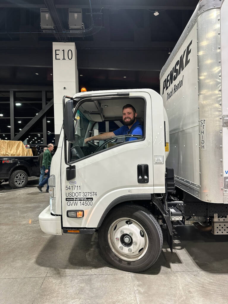 Young man with beard in blue shirt sitting in the cab of a white delivery truck with blue lettering inside a convention hall. 