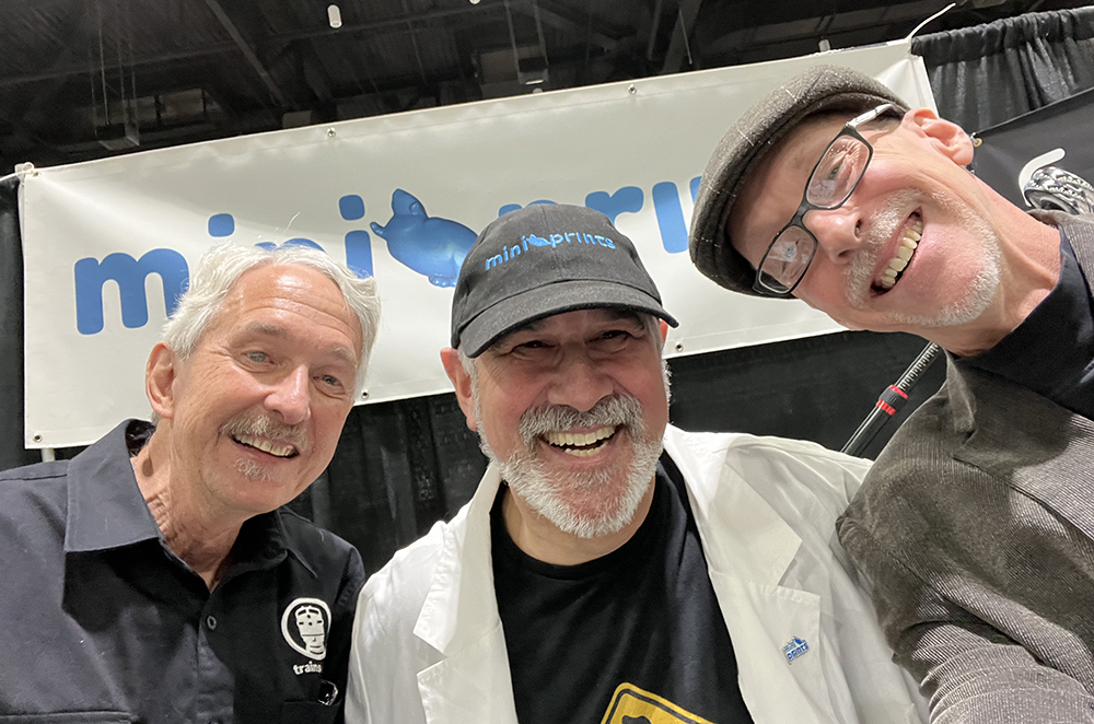 Three older men, one wearing a white coat and gray hat, one wearing glasses, a tan coat, and a grey hat, and one with gray hair and no coat or hat, all smiling while taking a selfie photo. 