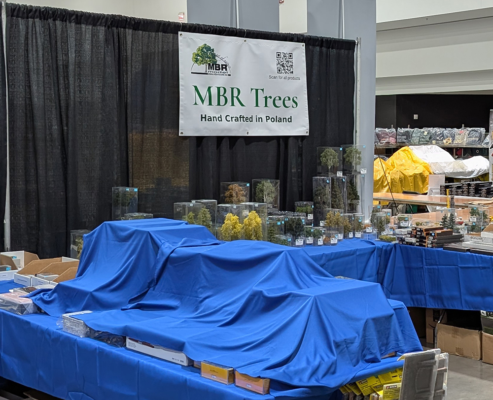 Trade show scene with blue drapery backdrop showing long tables with assorted model train products covered with blue clothes and a white sign with green lettering in front of assorted model trees in clear display boxes. 