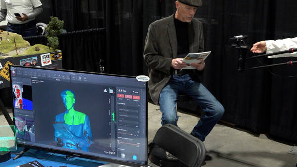 Older man with gray cap and tan coat holding a blue and white book while seated on a stool in front of a computer screen with a blue, green and gray computer-generated identical image of him.