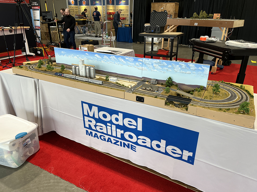 Highly detailed oval-shaped miniature railroad with green grass, blue sky, gray and silver track, and tiny houses resting on a large, white table with a blue banner in front of it. 