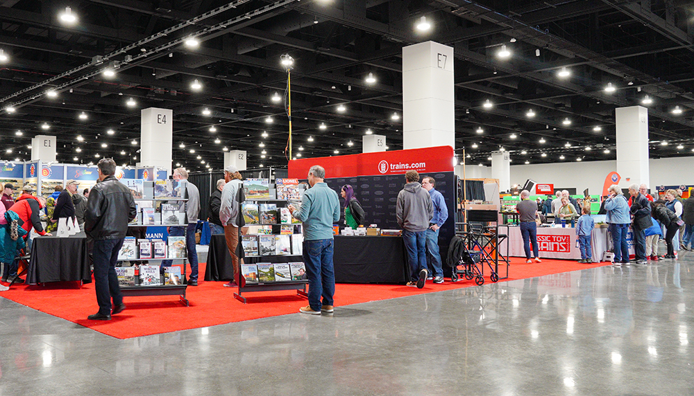 Trade show booth set on red carpet with black tables, red and black backdrop, and assorted gray bookshelves stocked with book all in a large, brightly lit convention hall filled with people. 