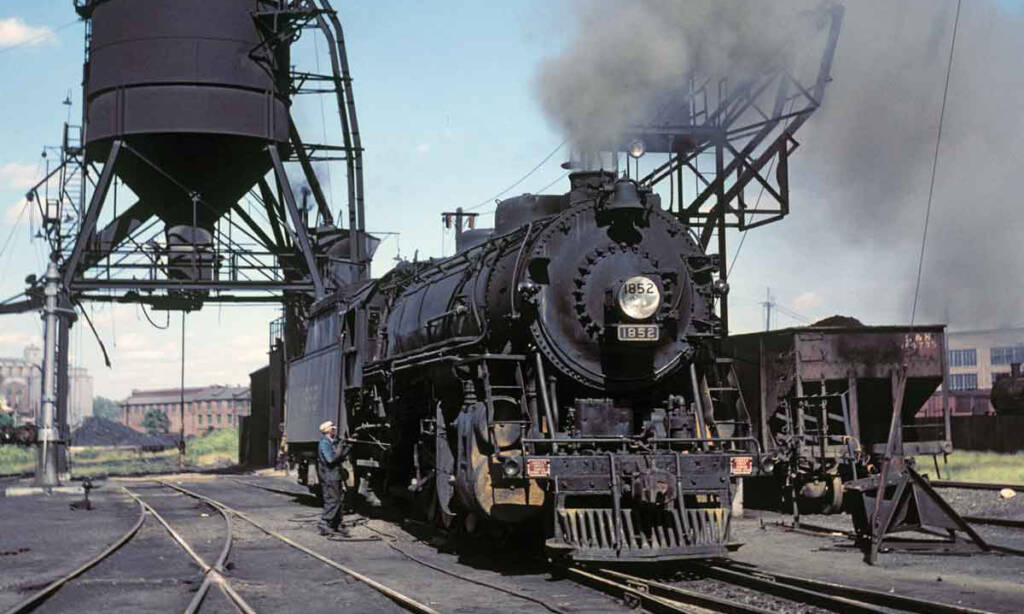 A color photograph of a black steam locomotive in a rail yard