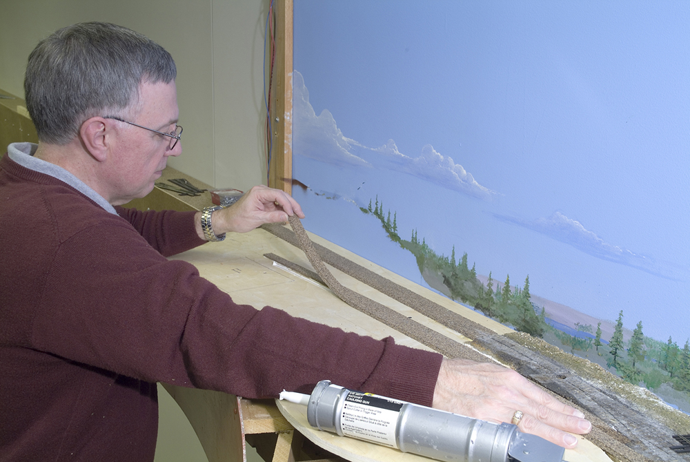 Color photo of man in dark red sweater attaching cork strip to model railroad.