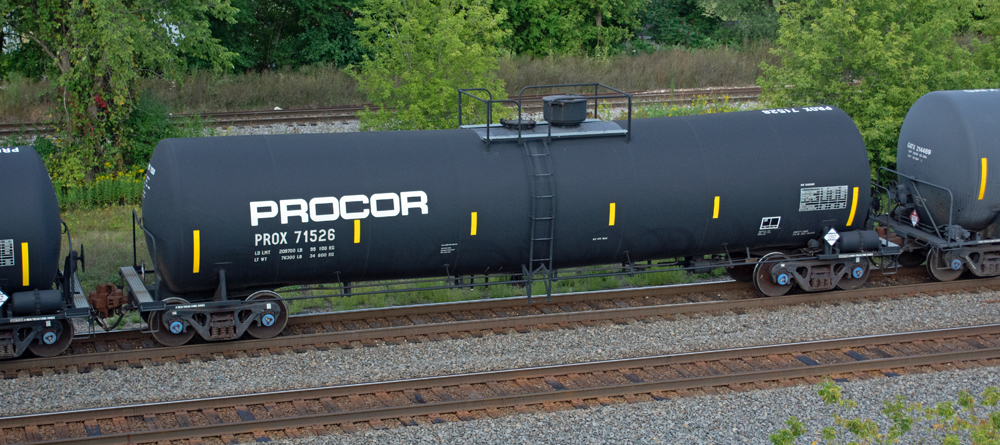 Color photo of black tank car passing through a wooded scene.