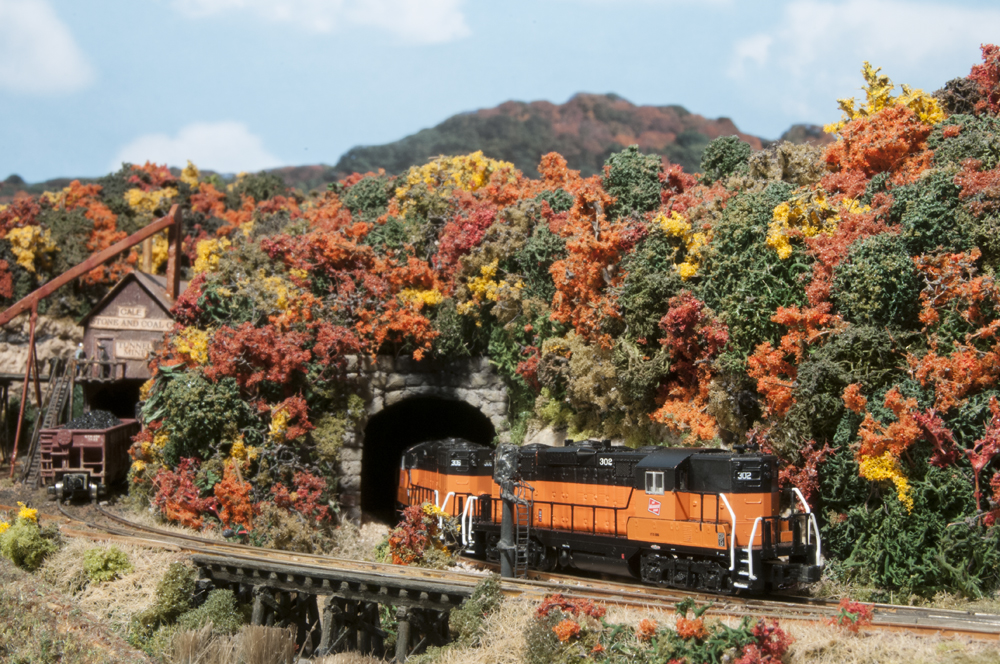 Color photo of Z scale Milwaukee Road EMD GP9s emerging from tunnel in a scene with fall foliage.