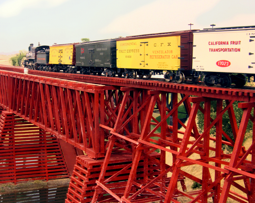 Color photo of steam locomotive and train passing over wood trestle and truss bridge.