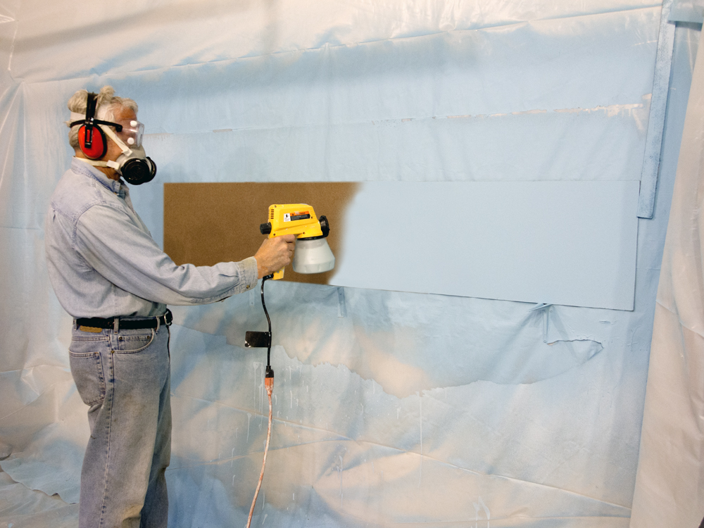 Color photo of man in denim shirt and blue jeans wearing personal protective gear painting tempered hardboard.