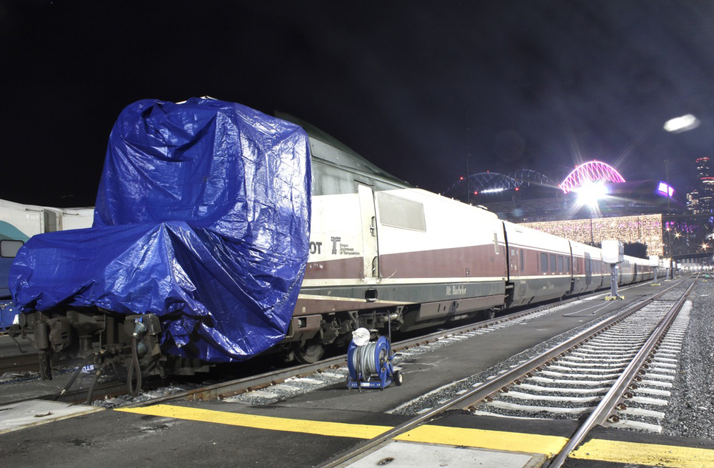 Permanently coupled passenger train with damaged nose covered by tarp in yard at night.