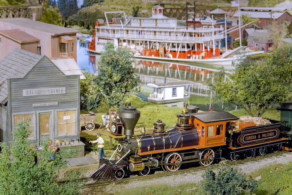 Model of a vintage steam locomotive passes by a 19th Century river town.