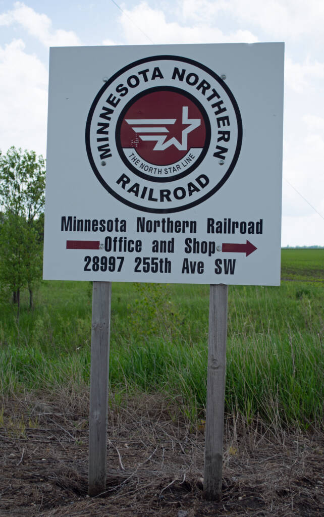 Color photo of white sign with maroon and black graphics attached to wood post in rural area.