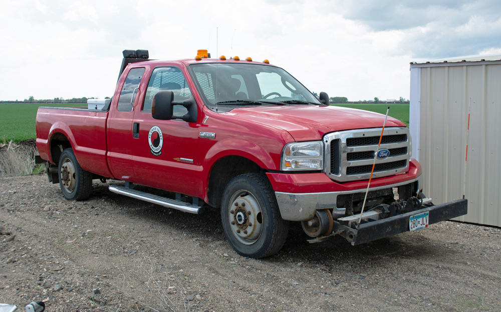 Color photo of red pickup truck with hi-rail vehicles next to tan shed.