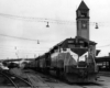 Square diesel locomotive with passenger train at station with tall tower