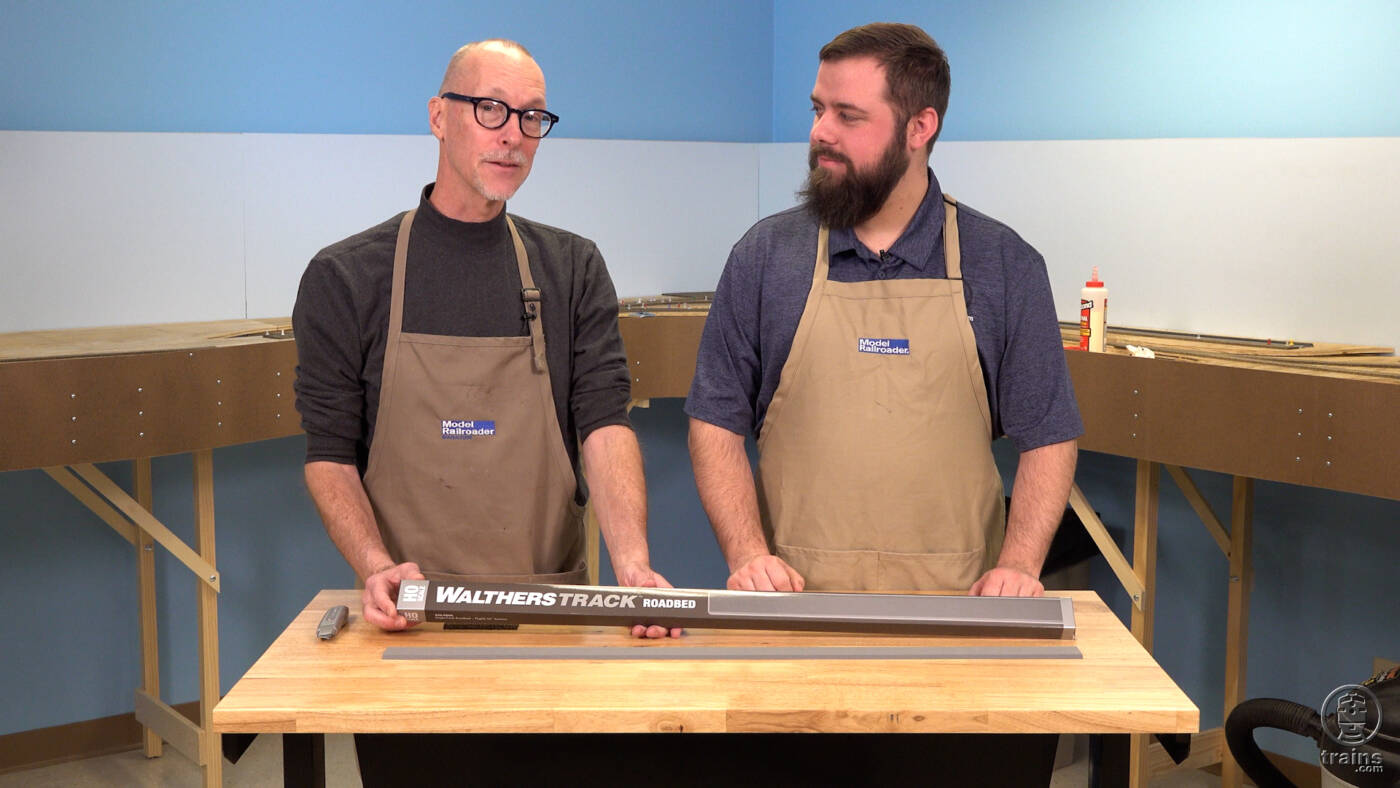 Two men in tan work aprons standing in front of workbench with model railroad in background