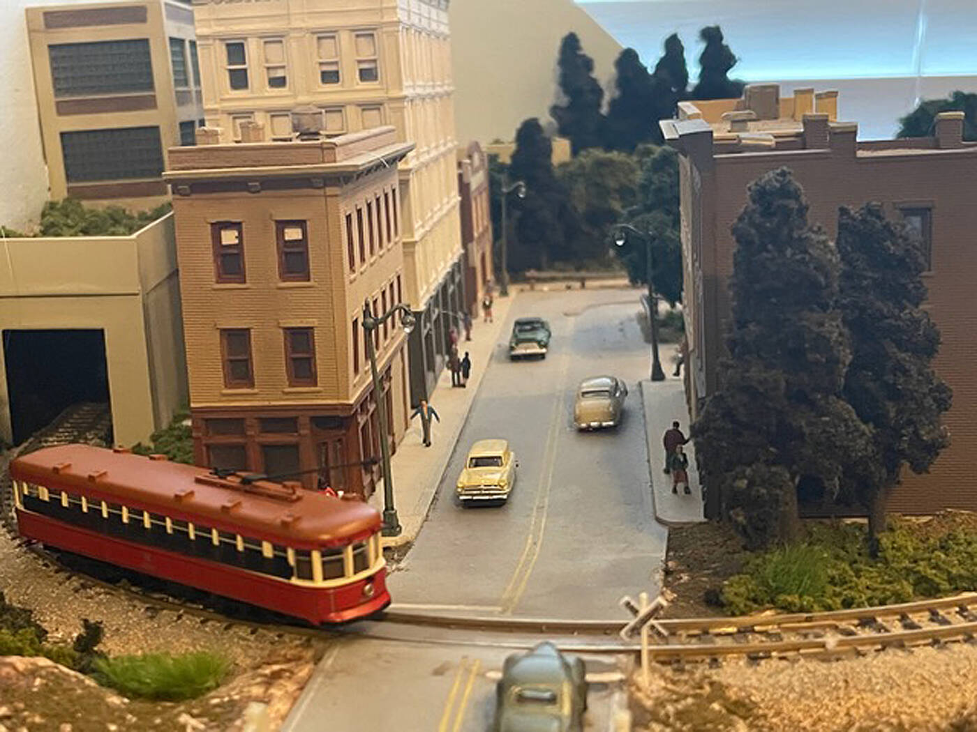 A close-up photo of a model railroad layout, focusing on a street level grade crossing with a red model trolley