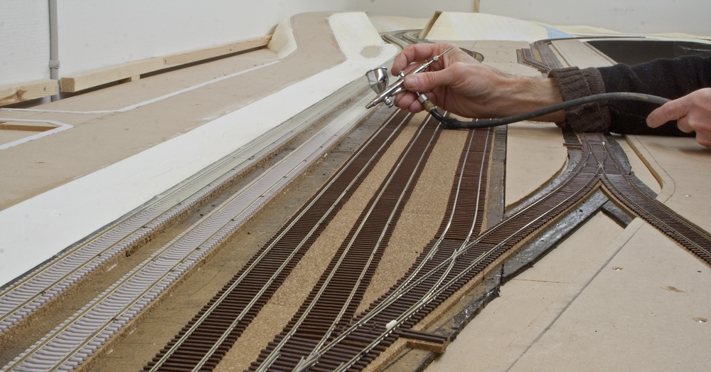 Photo showing man’s hand holding airbrush while painting model railroad track.