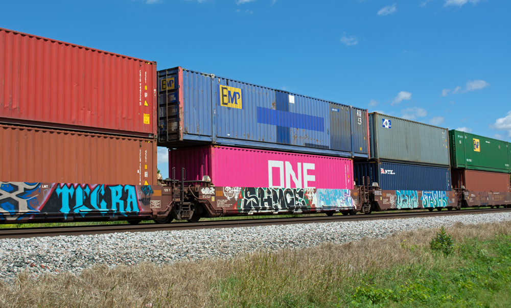 Color photo of articulated well car in mineral red paint covered with graffiti. Units are loaded with intermodal containers in various colors.