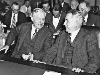 Two well-dressed men, smiling, sitting at a large table with other people in the background. Invisible Giants — inside the Van Sweringen brothers empire.